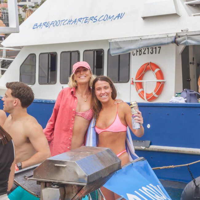 A group of people in swimsuits enjoy a party on a boat. Two women in front, smiling, stand near a grill; the boat behind them has a life preserver and a "barefootcharters.com.au" sign. Others socialize in the background.