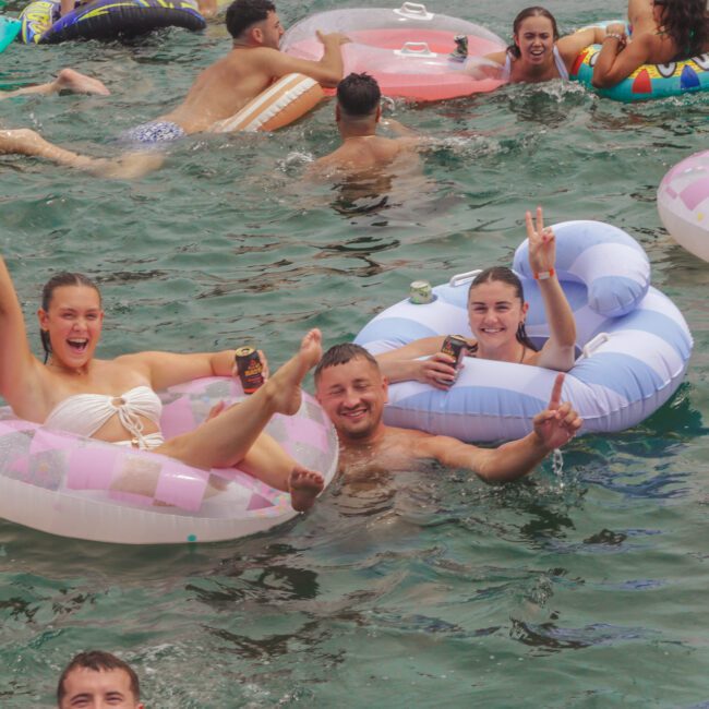 People relax and smile on colorful pool floats, holding drinks and making peace signs, while others swim around them in a lively outdoor water scene. The atmosphere looks festive and fun.