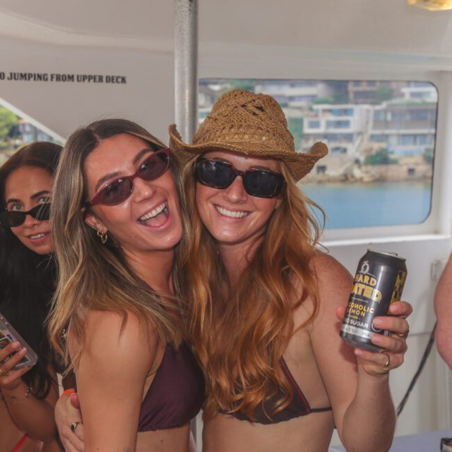 Two smiling women in swimsuits and sunglasses, one wearing a cowboy hat, pose together on a boat. One holds a drink can. Other people stand nearby, enjoying the sunny party atmosphere. Water and buildings are visible in the background.