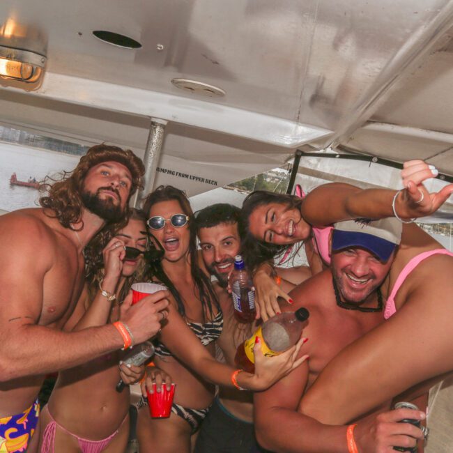 A group of young adults in swimsuits smile, laugh, and pose for the camera while partying on a boat. They are holding drinks and appear to be enjoying a lively, fun atmosphere.