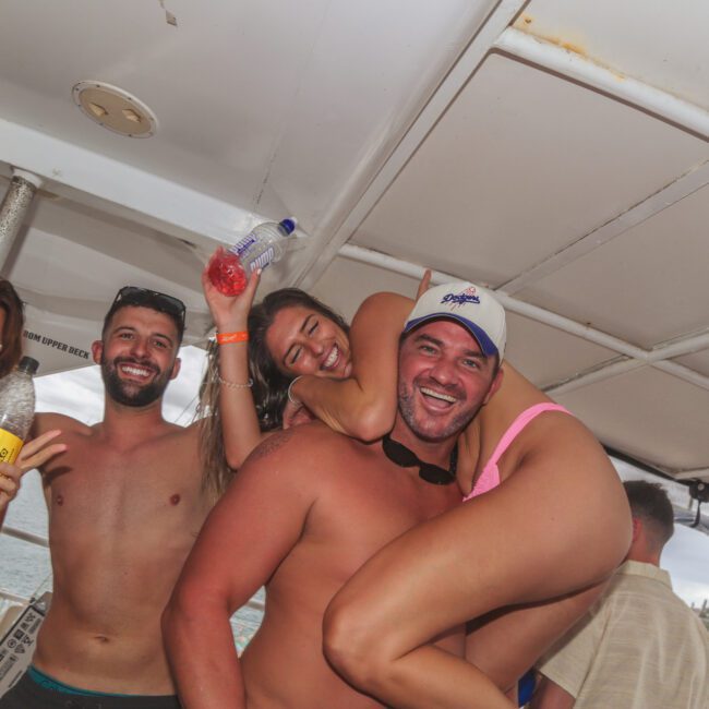 A group of young adults in swimsuits smile and pose for a lively photo on a boat. One woman playfully hugs a man from behind. Others hold drinks and wear sunglasses, enjoying a sunny day on the water.