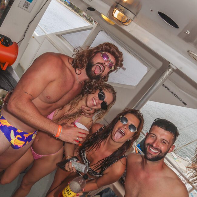 Four smiling adults in swimsuits pose together on a boat, holding drinks and wearing sunglasses. They appear to be enjoying a lively, fun atmosphere near the water, under a covered area of the boat.