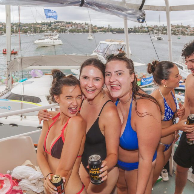A group of young adults in swimsuits smile and pose with drinks on a boat, with other boats and a city shoreline visible in the background. The atmosphere is lively and festive.