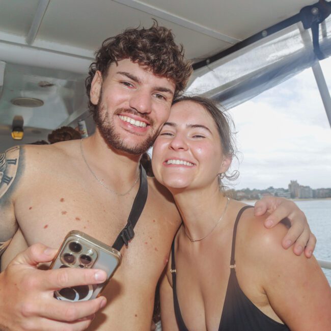 A smiling man and woman in swimsuits pose together on a boat, with water and city buildings visible in the background. The man holds a phone, and both appear happy and relaxed.