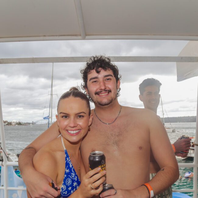 A young woman in a blue bikini and a shirtless young man smile at the camera on a boat. The man has his arm around the woman. Boats and people are visible on the water in the background under a cloudy sky.