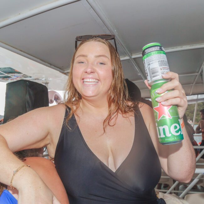 A smiling woman in a black swimsuit holds up a can of Heineken beer on a boat, surrounded by other people enjoying a social event.