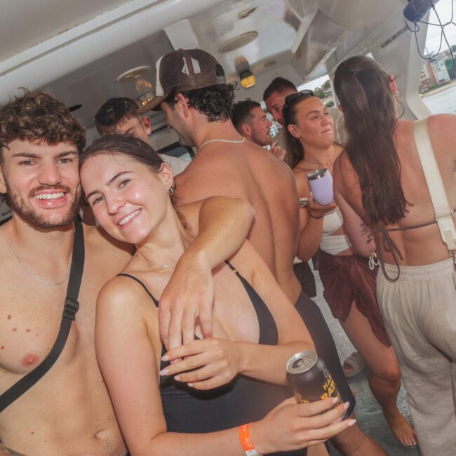 A group of young adults in swimwear enjoy a lively boat party. Two people in the foreground smile at the camera, one holding a drink. Others are chatting and dancing, with water and a shoreline visible through the windows.