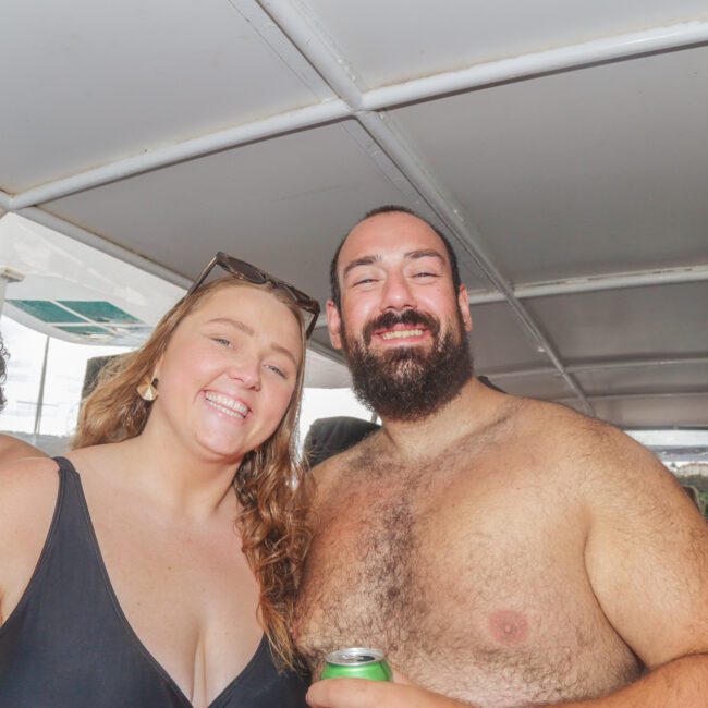 Two people in swimsuits smile and pose together on a boat. Other people are in the background, and water is visible outside. Both are holding drinks and appear to be enjoying a sunny day.