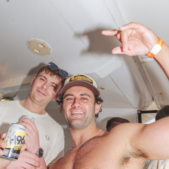 Two young men at a party; one is shirtless, wearing a cap, smiling and gesturing with his hand while holding a drink. The other man stands behind, smiling and wearing sunglasses on his head.
