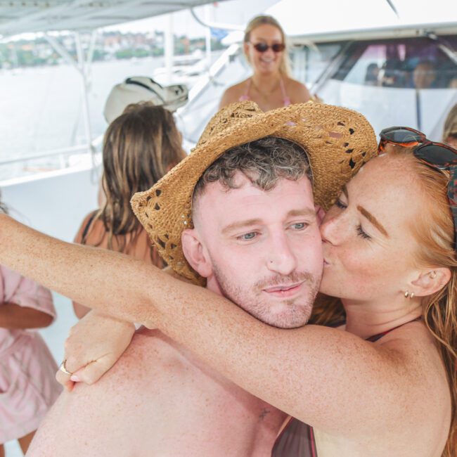 A woman in a swimsuit and straw hat kisses a shirtless man on the cheek at a lively boat party, with other people in swimsuits enjoying themselves in the background.