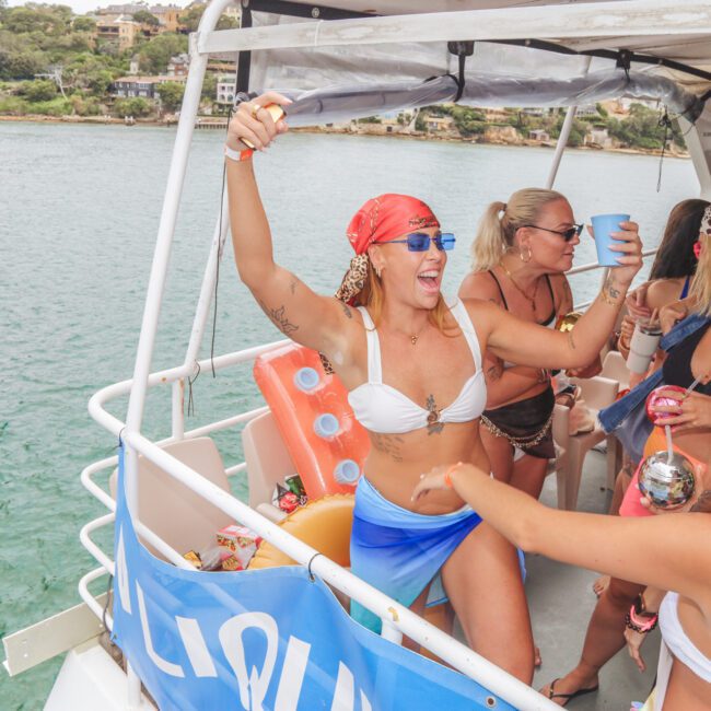 A group of people in swimwear dance and celebrate on a boat, holding drinks and smiling, while docked near the shore with trees and buildings visible in the background.