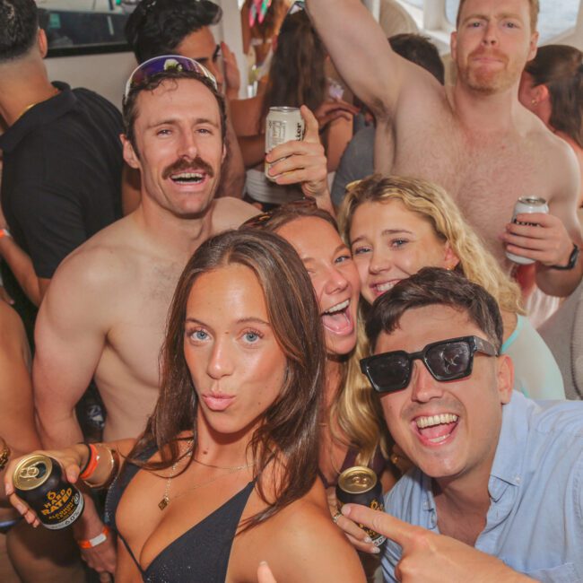 A group of young adults in swimwear smile and pose for a selfie at a lively boat party, holding drinks and wearing wristbands, with sunlight streaming through the windows.