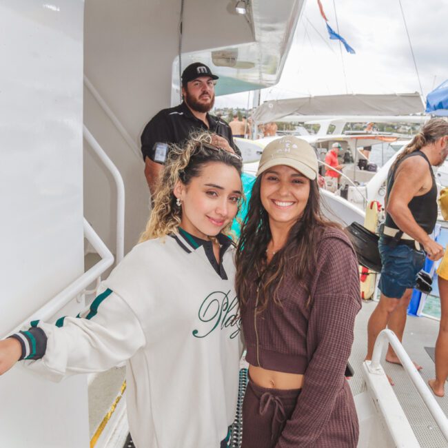 Two women pose and smile on the deck of a boat; one wears a cream sweater and shorts, the other a brown crop top and shorts set with a beige cap. Other people are in the background, enjoying the boat trip.