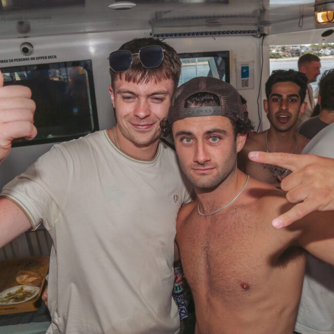 Two young men pose at a lively indoor party. One is shirtless and making a hand gesture, while the other smiles with his arm around him. People are socializing in the background. Bright lighting and a casual atmosphere.