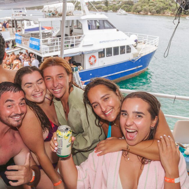 A group of five smiling young adults pose together on a boat, wearing swimsuits and towels, with one person holding a can. There are people and another boat in the water in the sunny background.