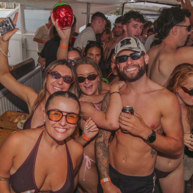 A group of people in swimsuits smile and pose for a photo at a crowded boat party, with drinks in hand and food on a table nearby. The atmosphere is lively and festive.