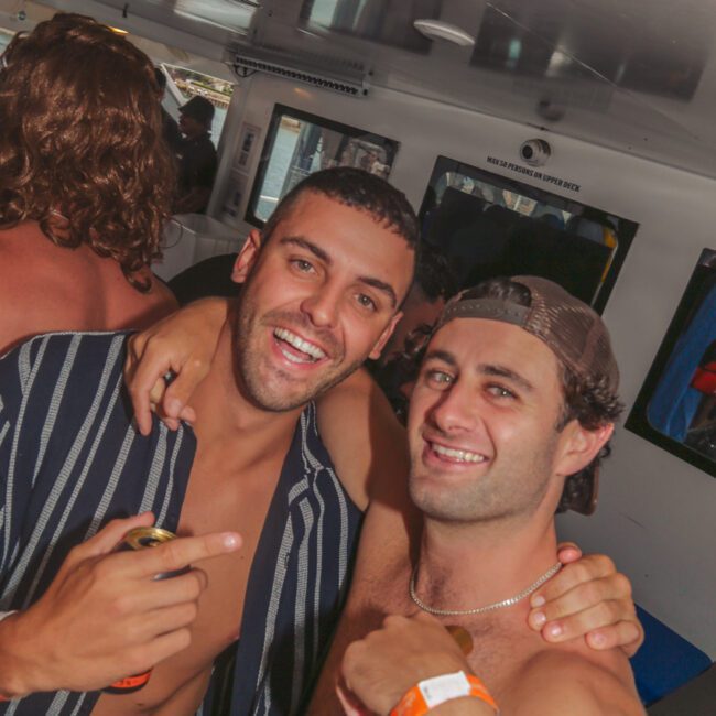 Two young men smile and pose together at a lively indoor party, one wearing a striped shirt and holding a drink, the other in a cap and wristband, surrounded by other people. A “Yacht Social” logo is visible in the corner.