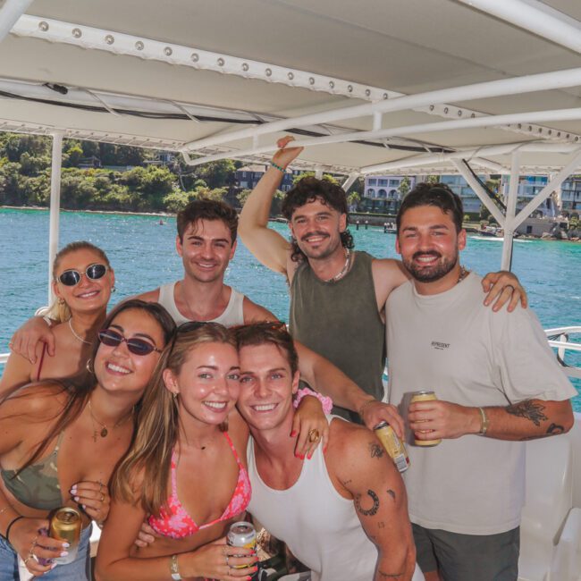 A group of six young adults, smiling and posing together on a boat. They are wearing casual summer clothes and sunglasses, holding drinks, with blue water and a green shoreline visible in the background.