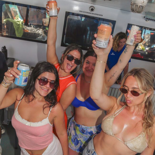 Four women in swimsuits and sunglasses cheerfully raise canned drinks while posing on a boat, smiling at the camera. Sunlight streams in through the windows, suggesting a lively, fun atmosphere.