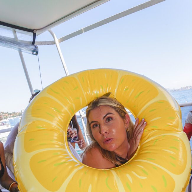 A woman poses playfully with her head through a yellow inflatable swim ring resembling a pineapple while on a boat, with water and shoreline visible in the background.