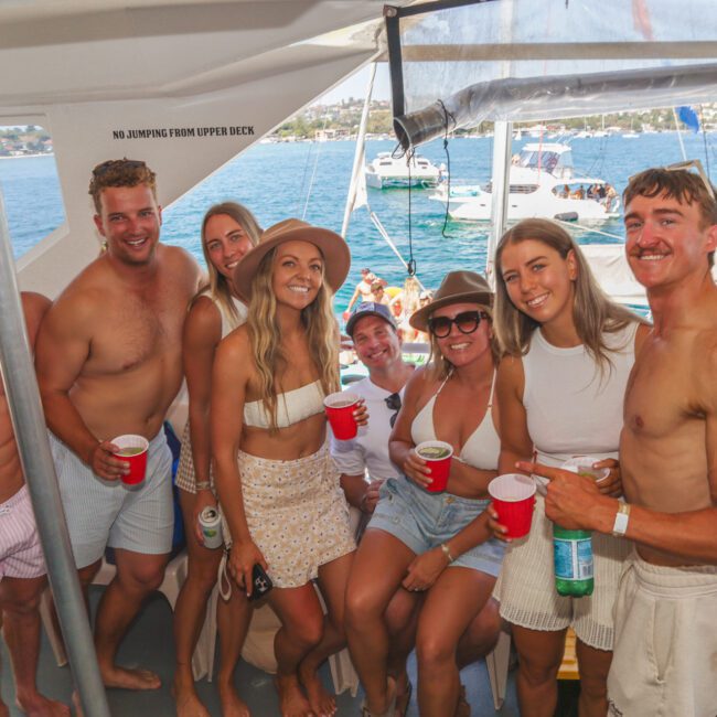 A group of smiling young adults in summer outfits pose together on a boat with drinks in hand. The sea and other boats are visible in the background on a sunny day.