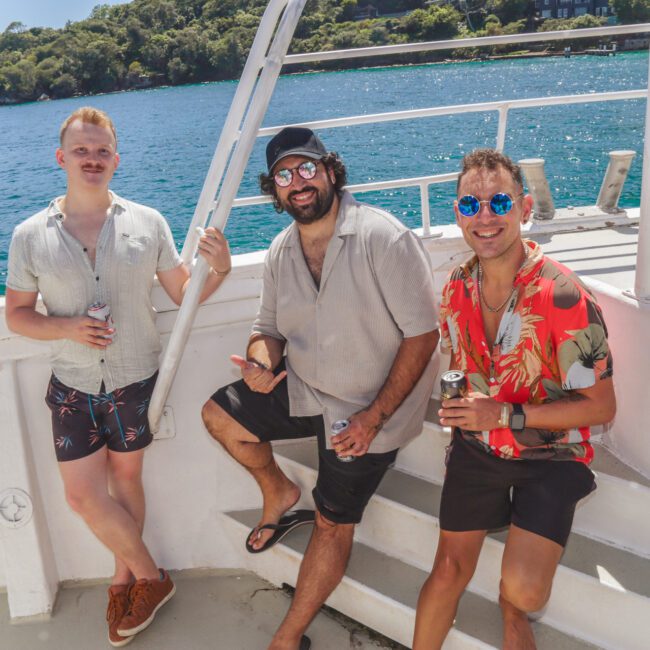 Three men pose and smile on a boat in sunny weather. They wear summer clothes and sunglasses, holding drinks, with blue water and green trees visible in the background.