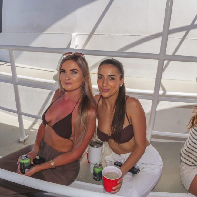 Two women in bikinis sit on a white bench on a boat, holding drinks and smiling at the camera. Sunlight casts shadows on the deck, and another person is partially visible beside them.