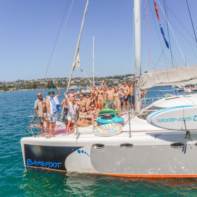 A group of people in swimsuits smile and pose on the deck of a white catamaran named "Barefoot," anchored on blue water with a cityscape and other boats in the background.