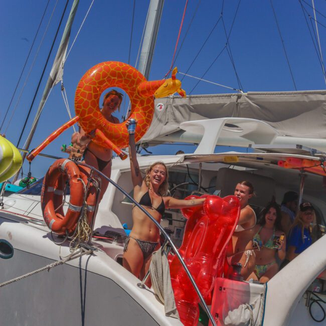 A group of people in swimsuits smiling and holding inflatable pool toys on the deck of a yacht under a sunny blue sky. Some are raising their arms and appear to be celebrating.
