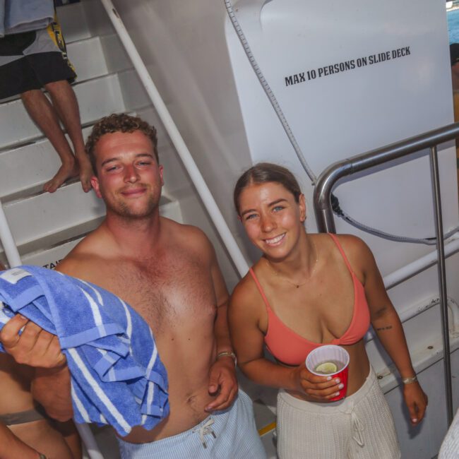 Three young adults in swimwear smile at the camera on a boat. One man holds a blue-striped towel, and a woman holds a red cup. A staircase and a sign saying “MAX 10 PERSONS ON SLIDE DECK” are visible in the background.