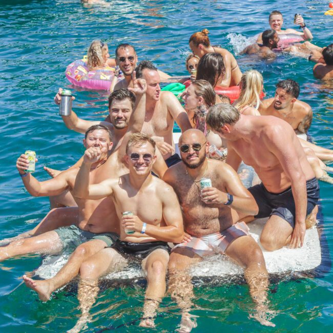 A group of people in swimsuits relax and smile on a floating platform in a sunny, blue body of water, holding drinks. Others float nearby on inflatable rafts, enjoying a lively summer party atmosphere.