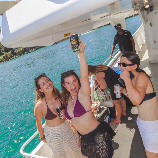 Three women in swimsuits smile and pose with drinks on the deck of a boat by blue water. One woman raises her can in the air. Other people and a crew member are in the background. It's sunny and festive.