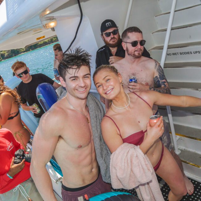 A group of young adults in swimsuits smile and pose on the deck of a boat in sunny weather, with water and trees in the background. Some are holding drinks, and others are standing on the stairs behind them.