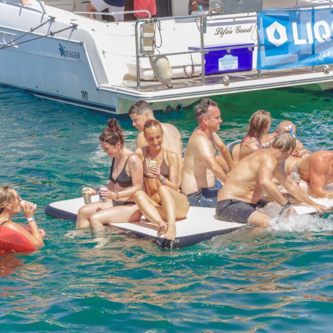 A group of people in swimsuits relax and socialize on a floating platform in clear blue water near a yacht, some holding drinks. One person lounges on an inflatable float nearby.