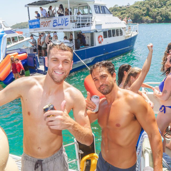 Two smiling men in swim trunks pose with drinks at a lively boat party. Other people socialize and dance on boats in the background on a sunny day, with clear blue water and green trees nearby.