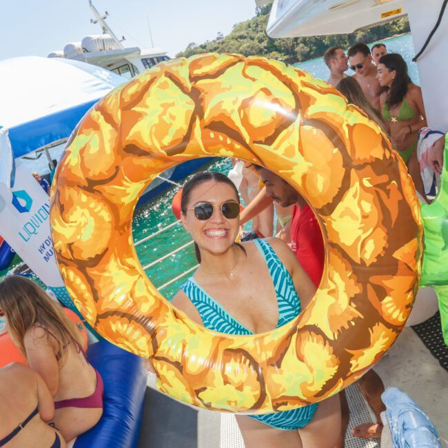 A woman in sunglasses and a striped swimsuit smiles while holding a pineapple-shaped float on a boat with people in swimsuits, inflatable toys, and blue water in the background.