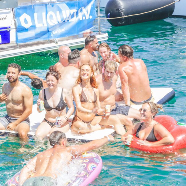 A group of people in swimsuits are smiling and relaxing on a floating platform and inflatable toys in the water near boats on a sunny day. Some are holding drinks and appear to be enjoying a lively, fun atmosphere.