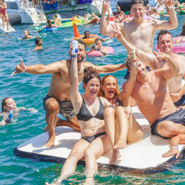 A group of people in swimwear smile, cheer, and pose on a floating mat in a crowded, lively blue water scene, with others swimming and lounging on inflatables and boats in the background.