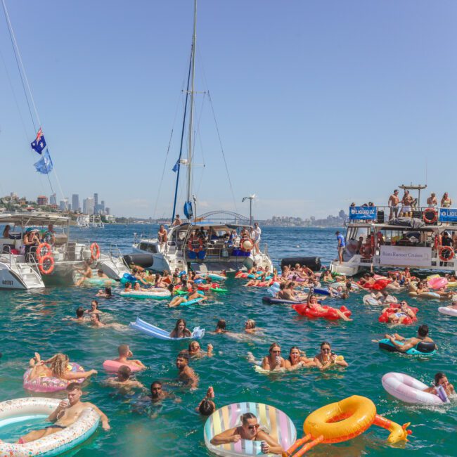 People on colorful inflatables and boats enjoy a crowded party in a sunny harbor, surrounded by water and city skyline in the background. The atmosphere is lively and festive.