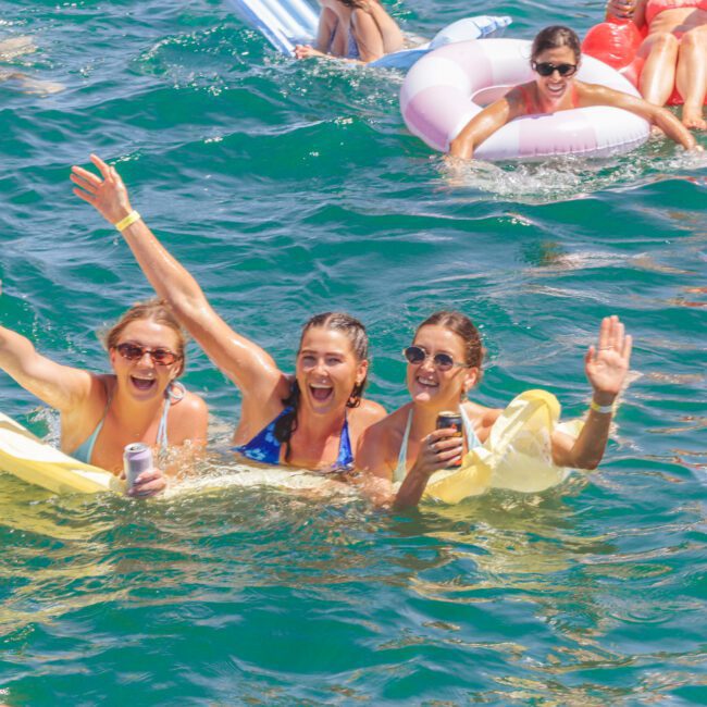 A group of people enjoying a sunny day on the water, some floating on inflatables and others swimming, with three women in the center smiling, cheering, and waving at the camera.