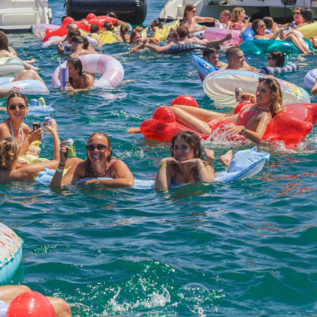A large group of people relax on colorful pool floats and inflatables in a crowded, sunny pool or ocean setting, enjoying a lively summer party near anchored boats.