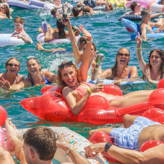 A group of young adults enjoy a sunny day in the water, smiling and laughing on colorful inflatable pool floats. The atmosphere is lively and festive, with many people relaxing and having fun in the blue water.