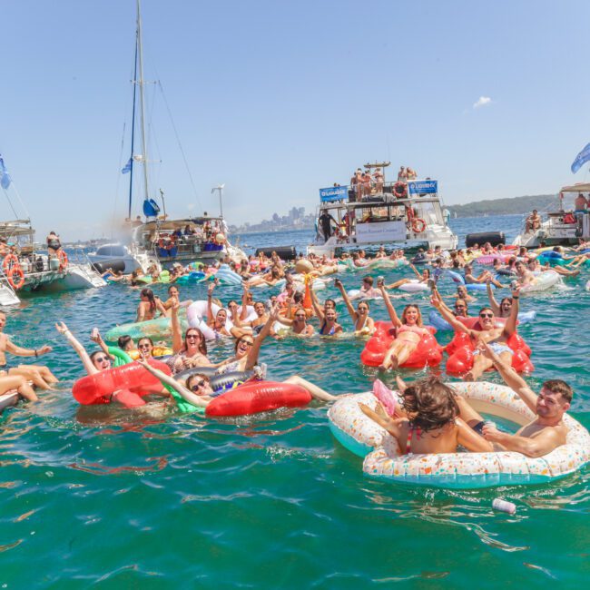 A lively group of people relax and party on colorful inflatables in the water, surrounded by several yachts and boats under a sunny sky. Many are smiling, waving, and enjoying the festive atmosphere.