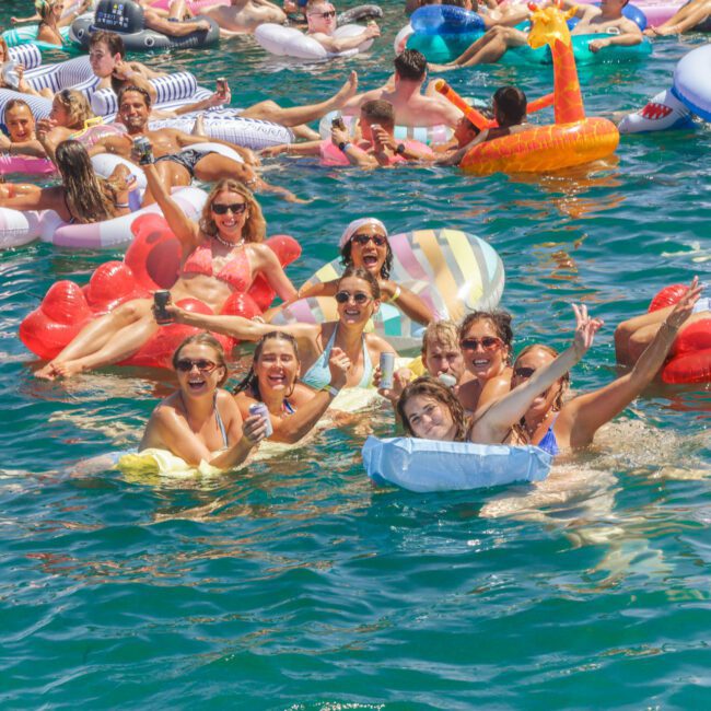 A large group of people on inflatable pool floats enjoy a sunny day on the water, laughing, smiling, and giving thumbs up. The scene is lively and festive, filled with bright colors and summer vibes.