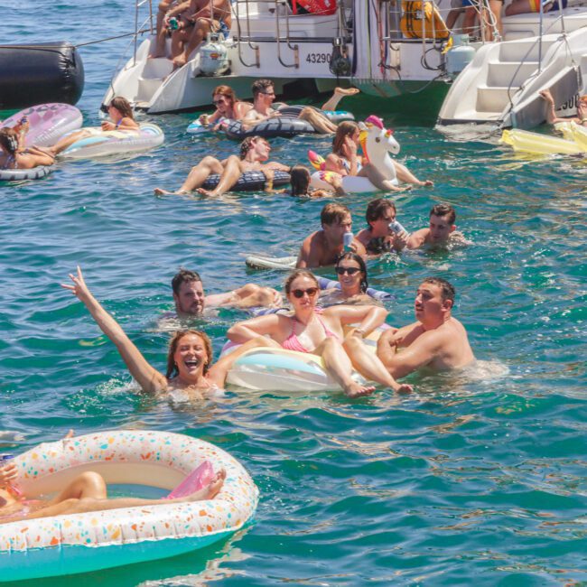A lively group of people enjoying a pool party in the ocean, floating on colorful inflatables near boats on a sunny day. Some are relaxing, others are swimming or celebrating with raised arms.