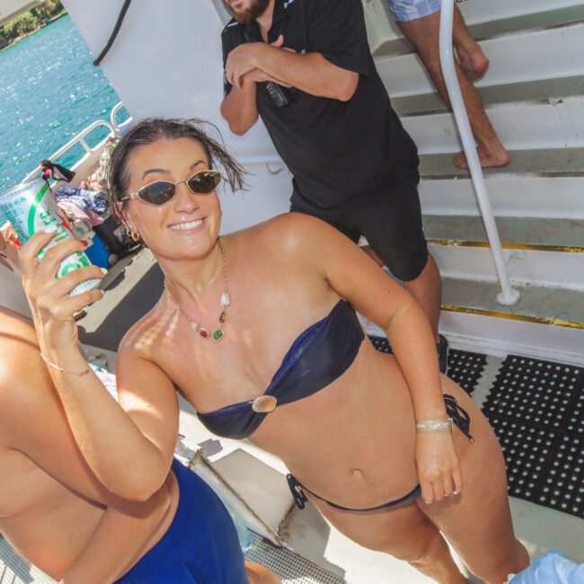 A woman in a blue bikini and sunglasses smiles while holding a can, standing on a boat near stairs. Other people are around, with water and greenery visible in the background. It is a sunny day.