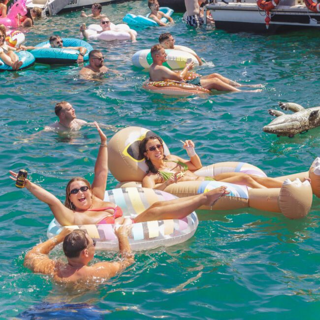 People relax and socialize on colorful pool floats in a sunlit, crowded lake. Two women in the foreground smile and gesture at the camera while lounging on inflatable floats. Others swim and float nearby.