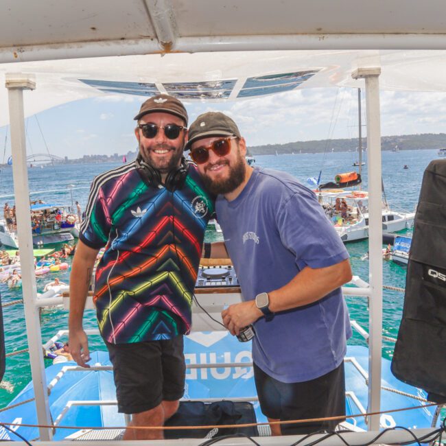 Two smiling men wearing sunglasses and hats stand at a DJ booth on a boat, surrounded by speakers. The background shows a lively scene with boats, people, and a city skyline under a sunny sky.