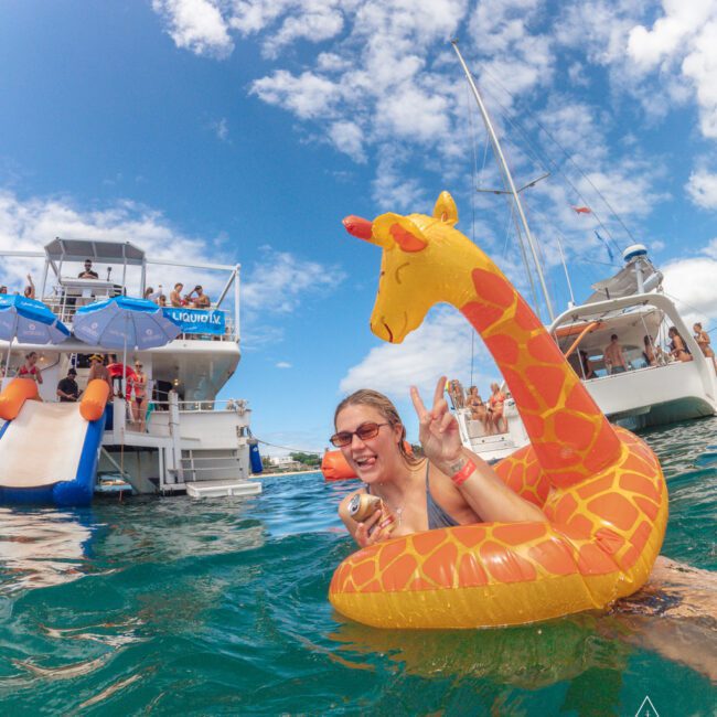 A woman in sunglasses smiles and flashes a peace sign while floating in the water with an inflatable giraffe. Behind her are boats, people, and a water slide under a bright blue sky with clouds.