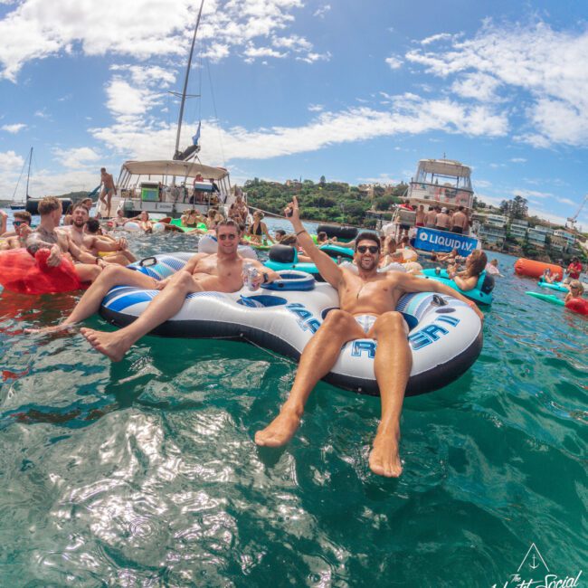 A group of people relax on large inflatable floats in clear blue water, smiling and celebrating near anchored boats under a partly sunny sky. Some wear sunglasses and swimsuits, enjoying a lively, festive atmosphere.